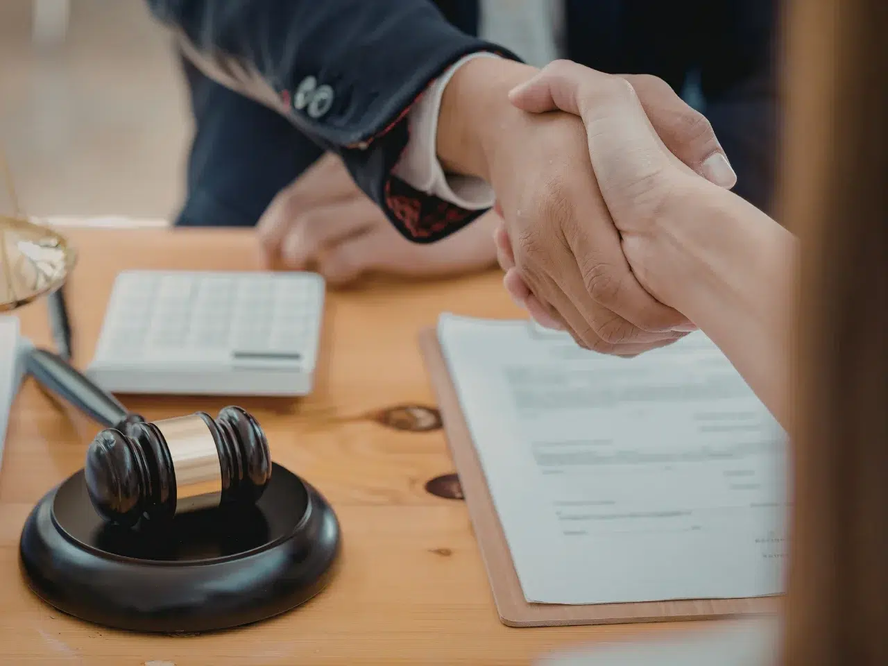 Two people shake hands over a desk with legal documents, a gavel, and a calculator, suggesting a legal agreement or settlement.