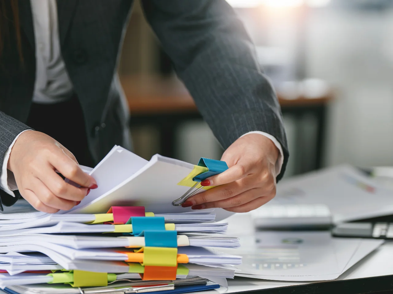 A person in a suit organizing stacks of documents with colorful paper clips and sticky notes on a cluttered desk in an office setting.