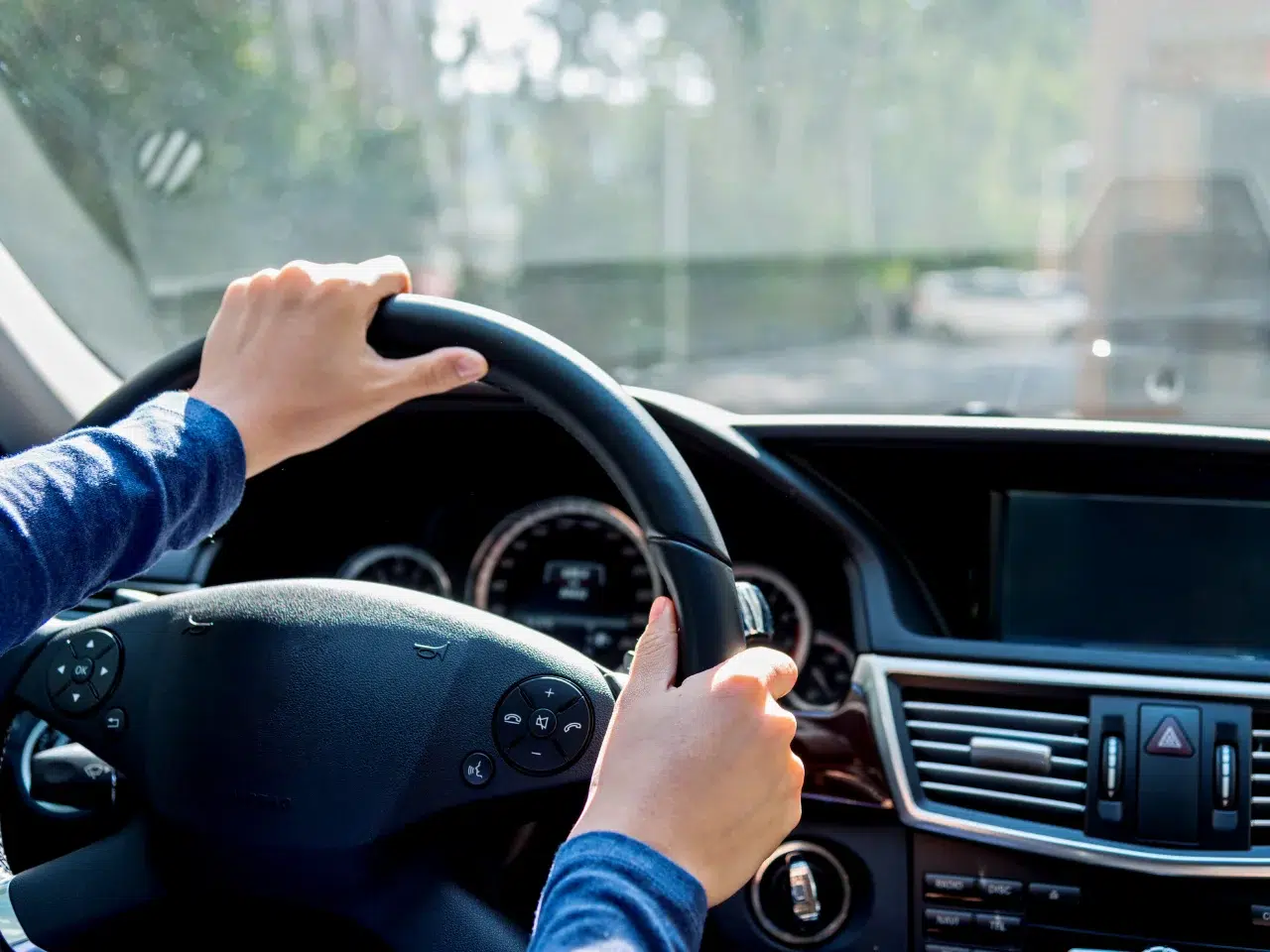 Close-up of a person’s hands on a car steering wheel, with the dashboard and controls visible, driving in daylight with a blurred view of the road and trees outside the windshield.