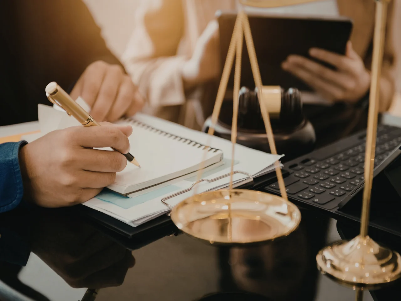 Two people sit at a desk with legal documents, a keyboard, and a tablet. One person writes in a notebook while the other holds a tablet. A gavel and scales of justice are visible in the foreground.