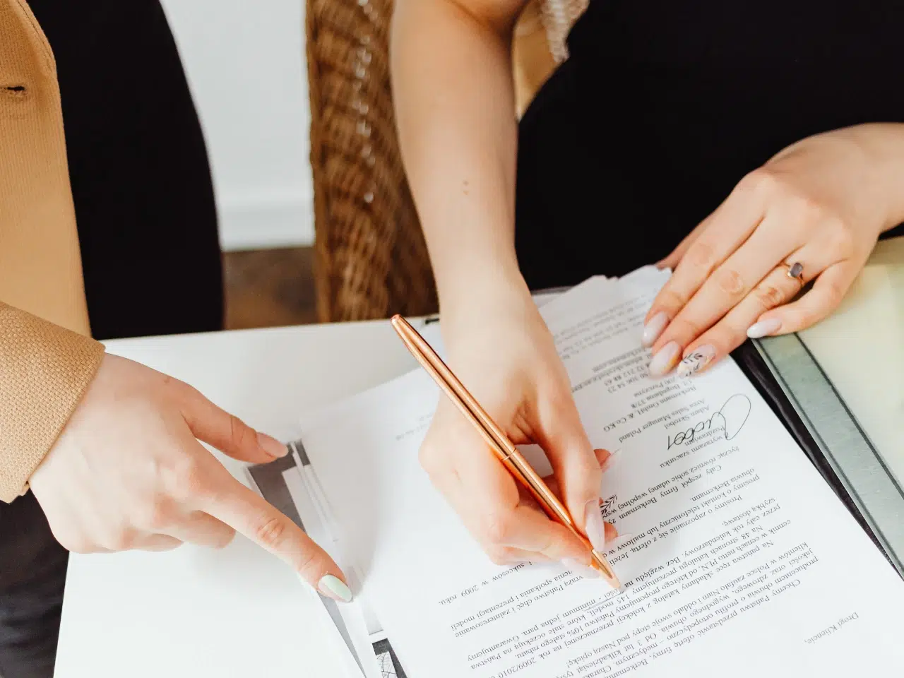 Two people are reviewing and signing documents on a desk; one person is holding a pen and writing, while the other is pointing at the papers. Only their hands and part of their arms are visible.