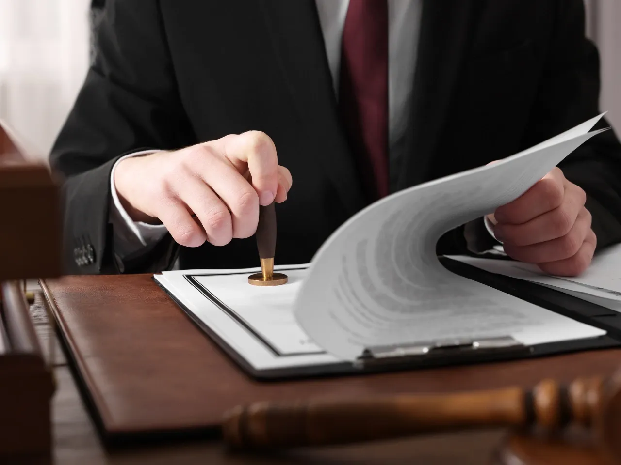 A person in a suit uses a wooden stamp on a document at a desk, with a gavel and more papers visible, suggesting a legal or official setting.