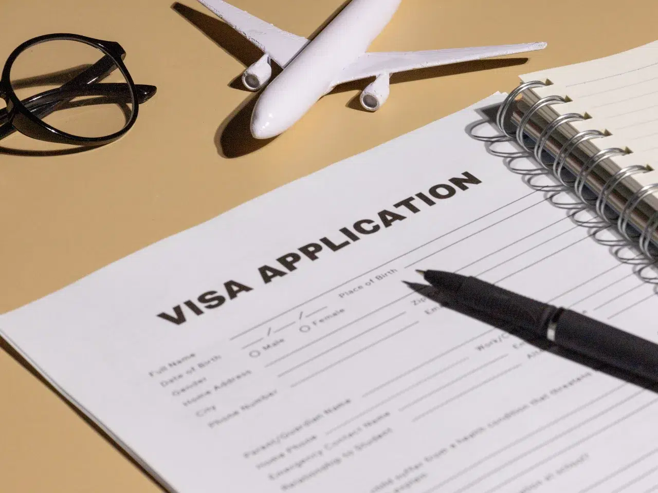 A visa application form on a desk with a black pen, spiral notebook, black eyeglasses, and a small model airplane, suggesting travel or immigration planning.