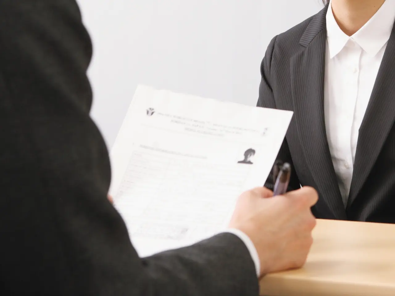 Two people in business suits sit at a table. One holds a pen and a printed resume with a small photo, suggesting a job interview or professional meeting. Only their hands and torsos are visible.