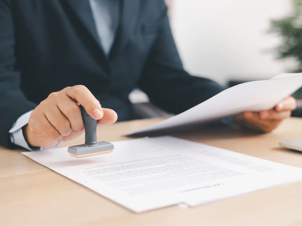 A person in a suit holds a document in one hand and presses a stamp onto a paper on a wooden desk, suggesting document approval or authorization.