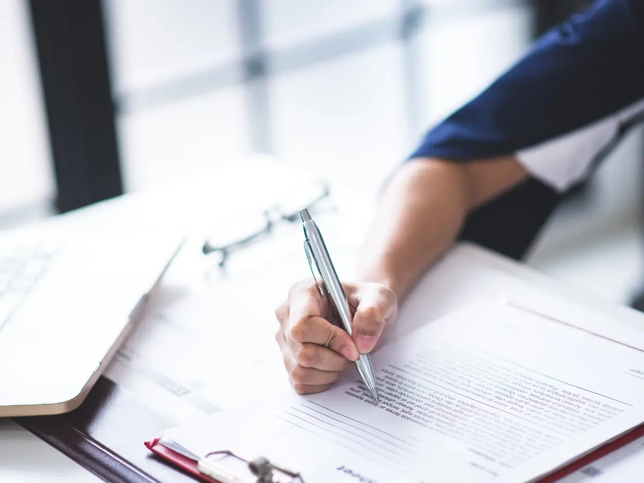 A person is signing a document with a pen at a desk. Nearby are a laptop, a pair of glasses, and additional papers with a red clipboard. The scene is well-lit, suggesting a professional or office environment.