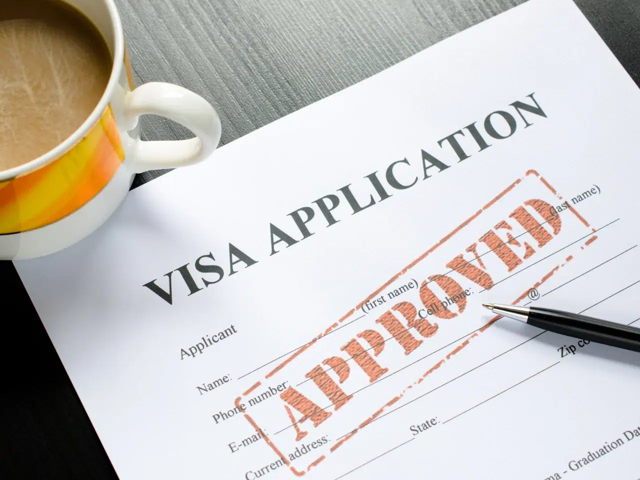 A visa application form on a table with a large red APPROVED stamp across it. A pen rests on the form and a cup of coffee sits nearby.