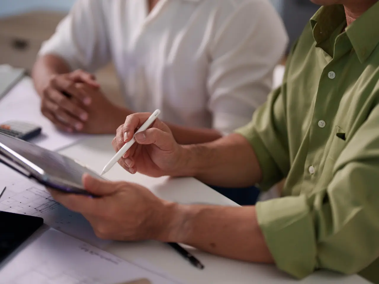 Two people sit at a table; one holds a stylus and a tablet, while the other sits with hands clasped. The focus is on their hands and the tablet, suggesting a discussion or collaboration.