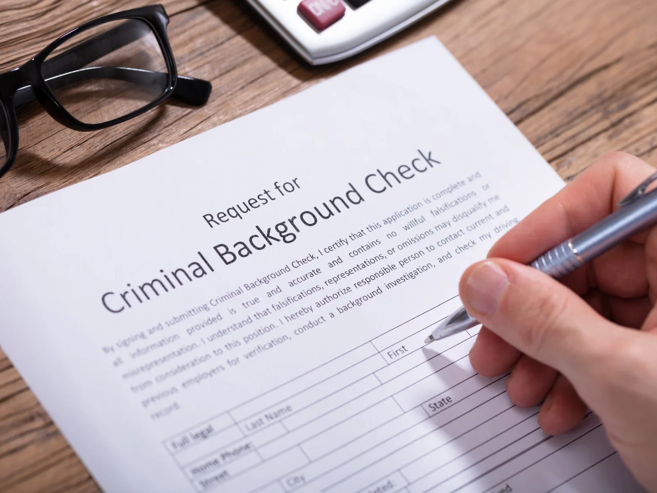 A hand fills out a Request for Criminal Background Check form on a wooden desk, with a pair of glasses and a calculator nearby.