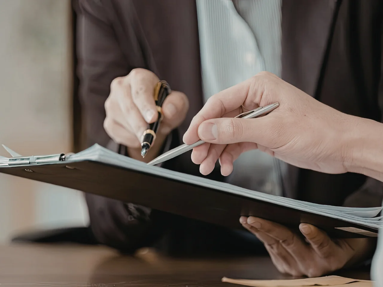 Two people holding pens, reviewing and discussing documents on a clipboard. One person points at the papers while the other gestures with a pen, suggesting a collaborative or business setting.