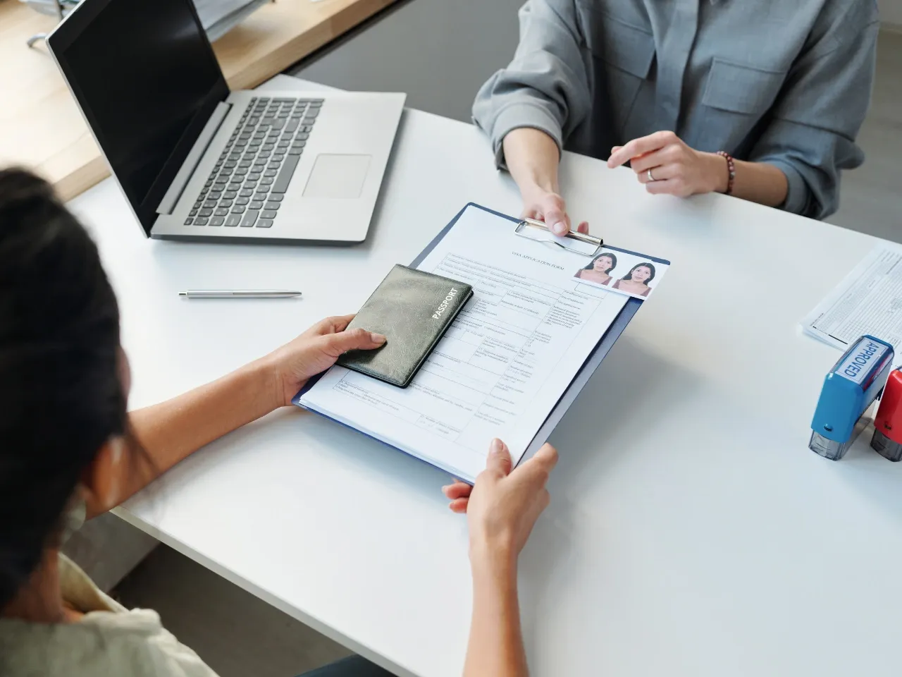 Two people sit across a desk from each other. One hands over a resume with photos and a passport. A laptop, pen, and office supplies are on the white desk, suggesting a job interview or document review.