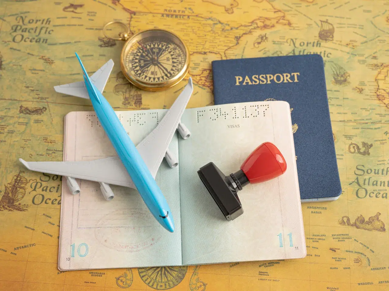 A blue toy airplane, a compass, a red stamp, and a blue passport rest on top of an open passport with visa stamps, all placed on a vintage world map background.