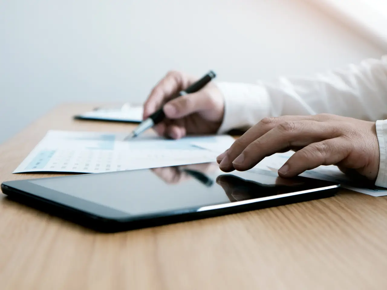 A person’s hands are visible on a desk, one holding a pen and writing on paper, the other resting near a tablet device. The scene suggests working with documents and digital technology.