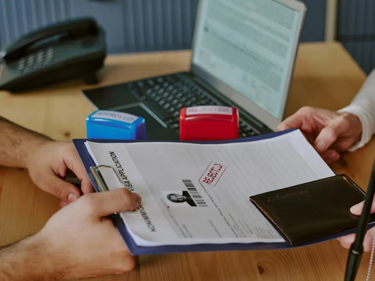 Two people exchange a clipboard holding a document stamped REJECTED, with a closed passport and pen visible. A laptop and two stamps—one blue, one red—are on the wooden desk in the background.