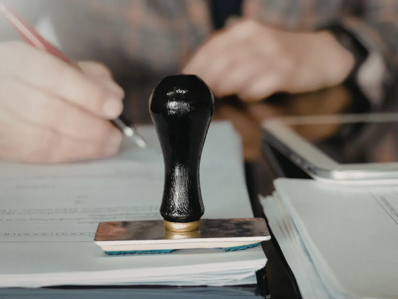 A close-up of a black rubber stamp resting on paperwork, with a person holding a pen and writing on documents in the background. A tablet and more papers are on the table.