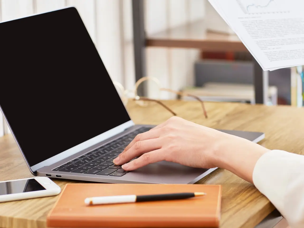 A person using a laptop on a wooden desk, with a smartphone, a closed notebook, and a pen nearby. The laptop screen is blank, and light papers are visible in the background.