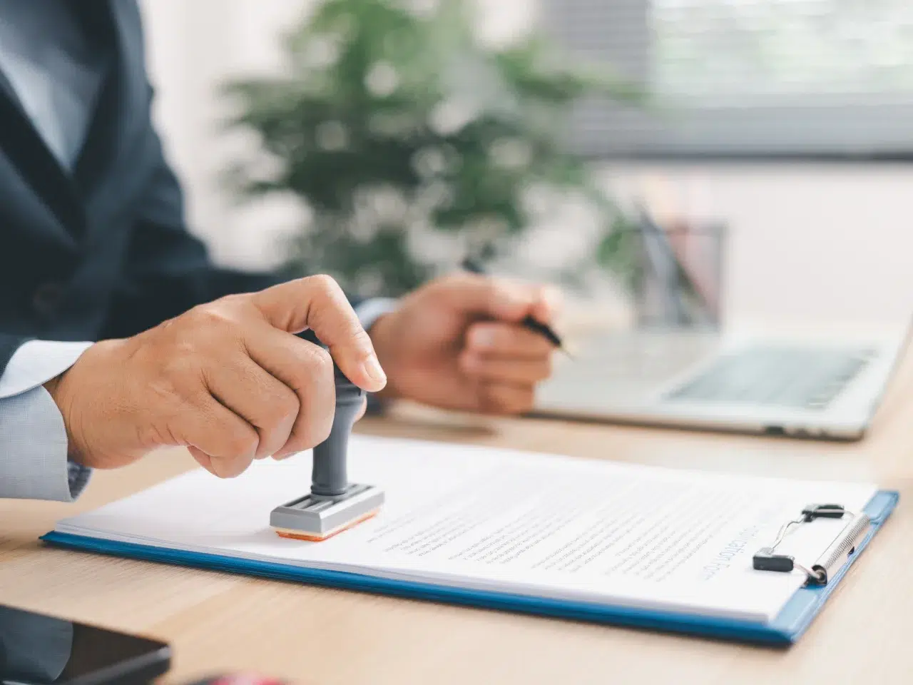 A person presses a stamp onto a document on a clipboard at a desk, with a laptop and a pen in hand. The background is blurred with a plant visible.