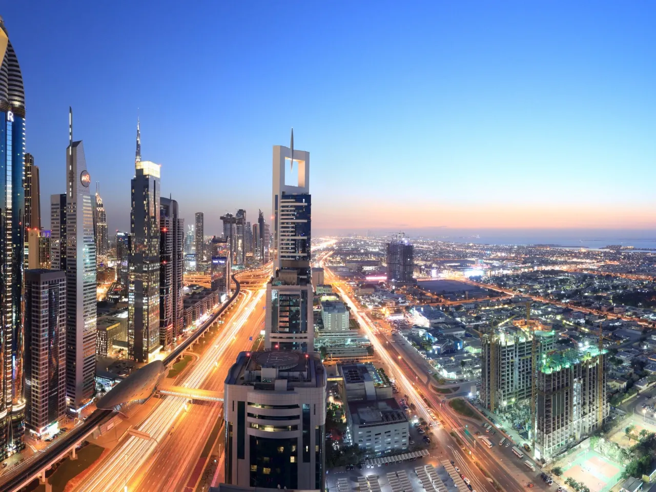 A cityscape of Dubai at dusk, featuring tall modern skyscrapers, illuminated roads with streaks of car lights, and a clear blue sky transitioning to orange near the horizon.
