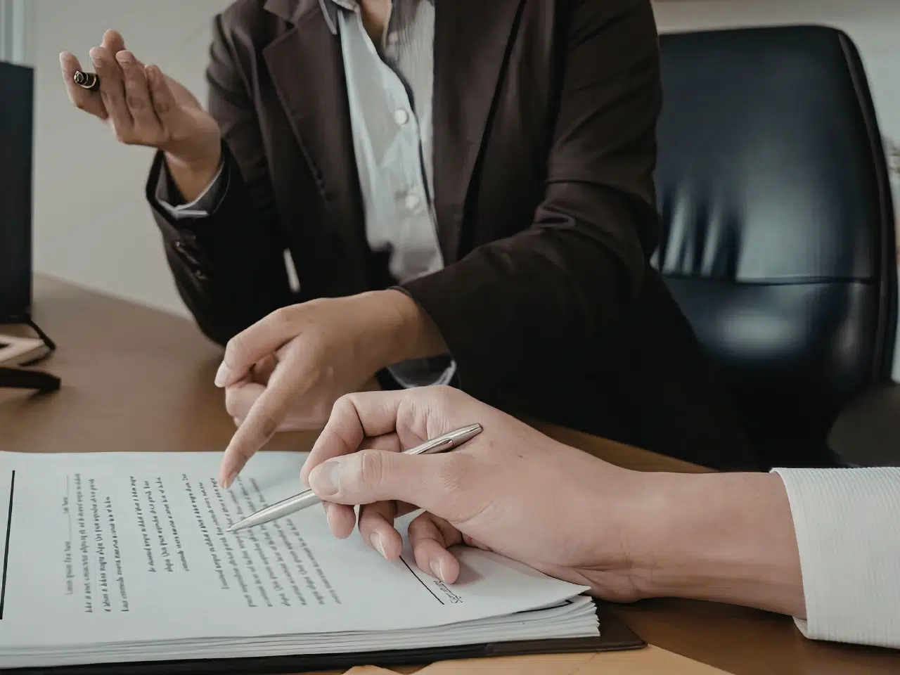 Two people in business attire sit at a desk discussing a document. One person points to text on the paper while the other holds a pen, ready to sign or make notes. The focus is on their hands and the document.