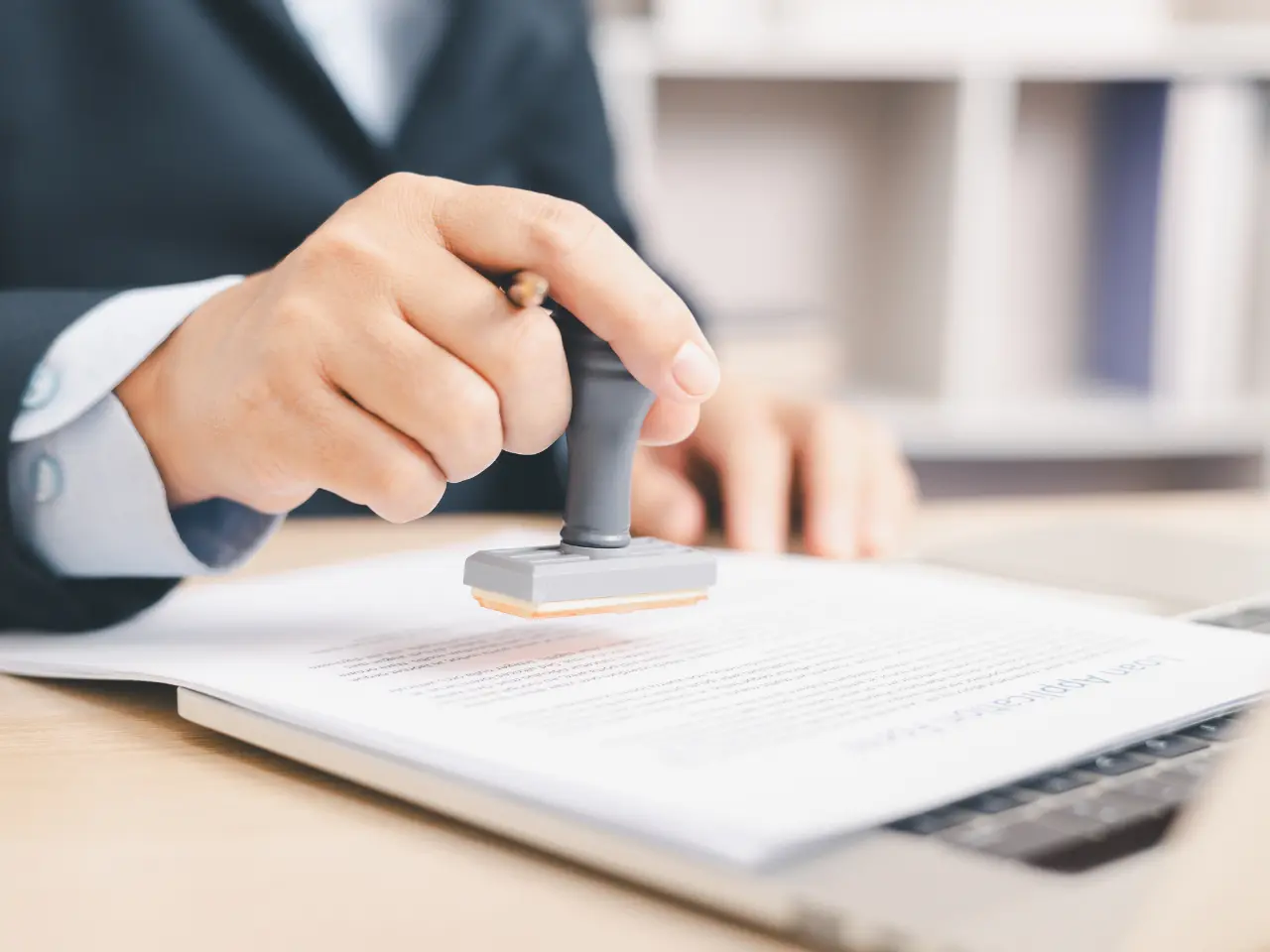 A person in business attire stamps a document placed on a desk, with a laptop partially visible nearby. The scene suggests approval or official validation of paperwork in a professional setting.