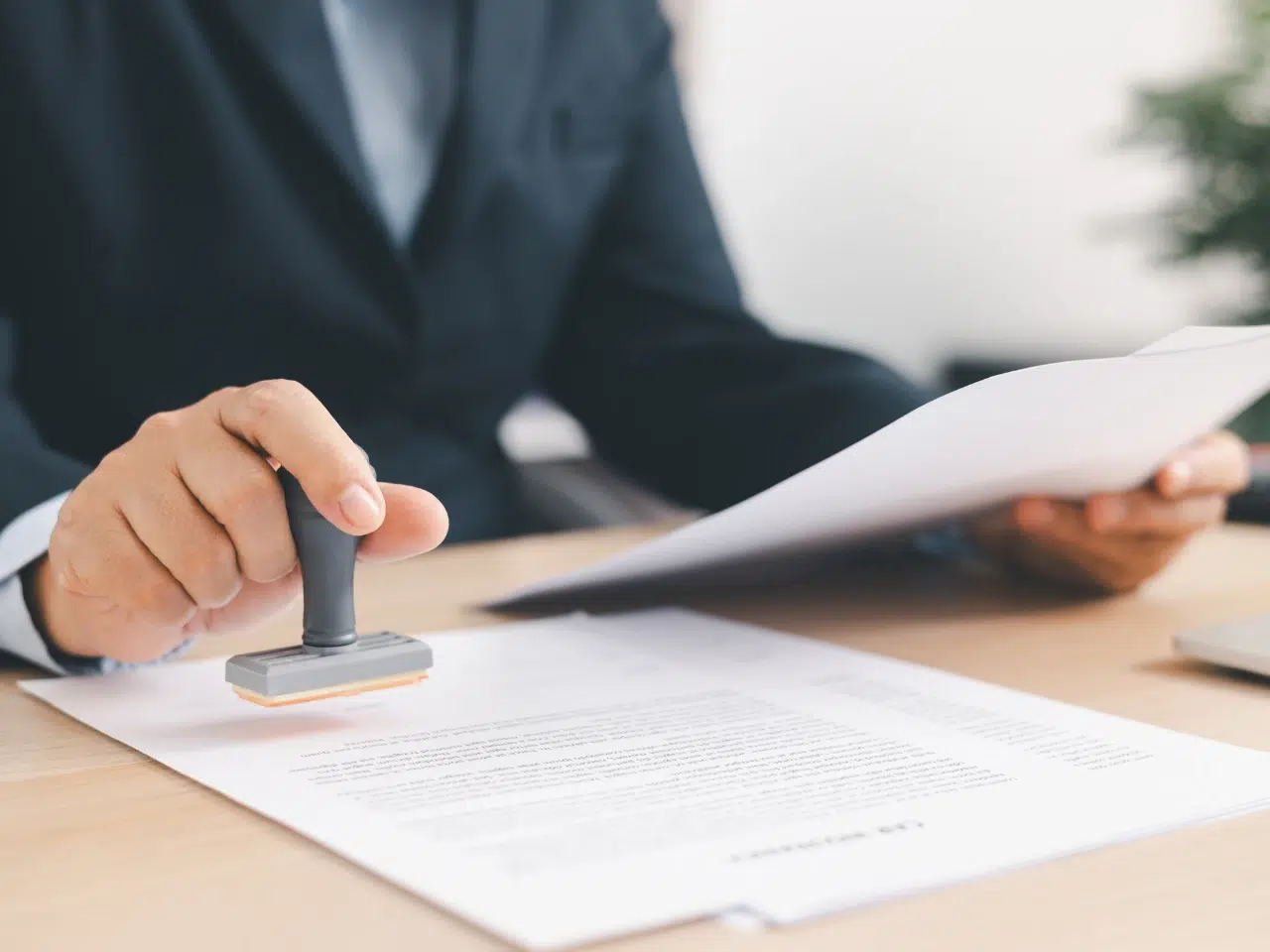 A person in a suit holds papers in one hand and uses a rubber stamp to approve or sign a document on a desk.