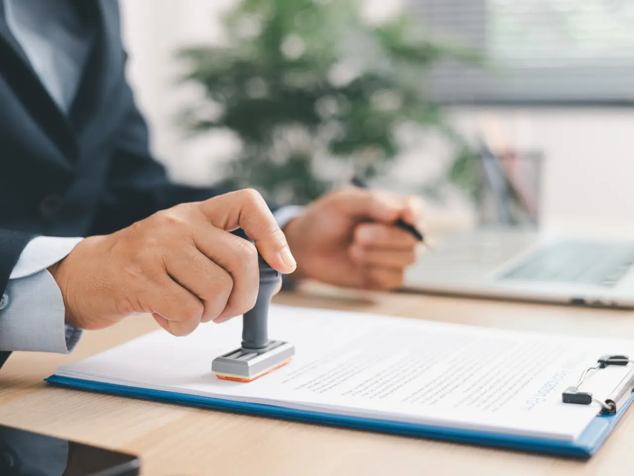 A person in business attire stamps an official document on a clipboard at a desk, with a laptop and other office supplies nearby.