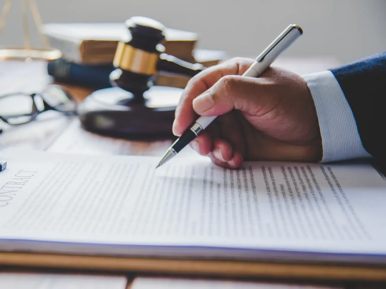 A person signing a contract document with a pen on a desk, with a judge’s gavel and legal books in the background.