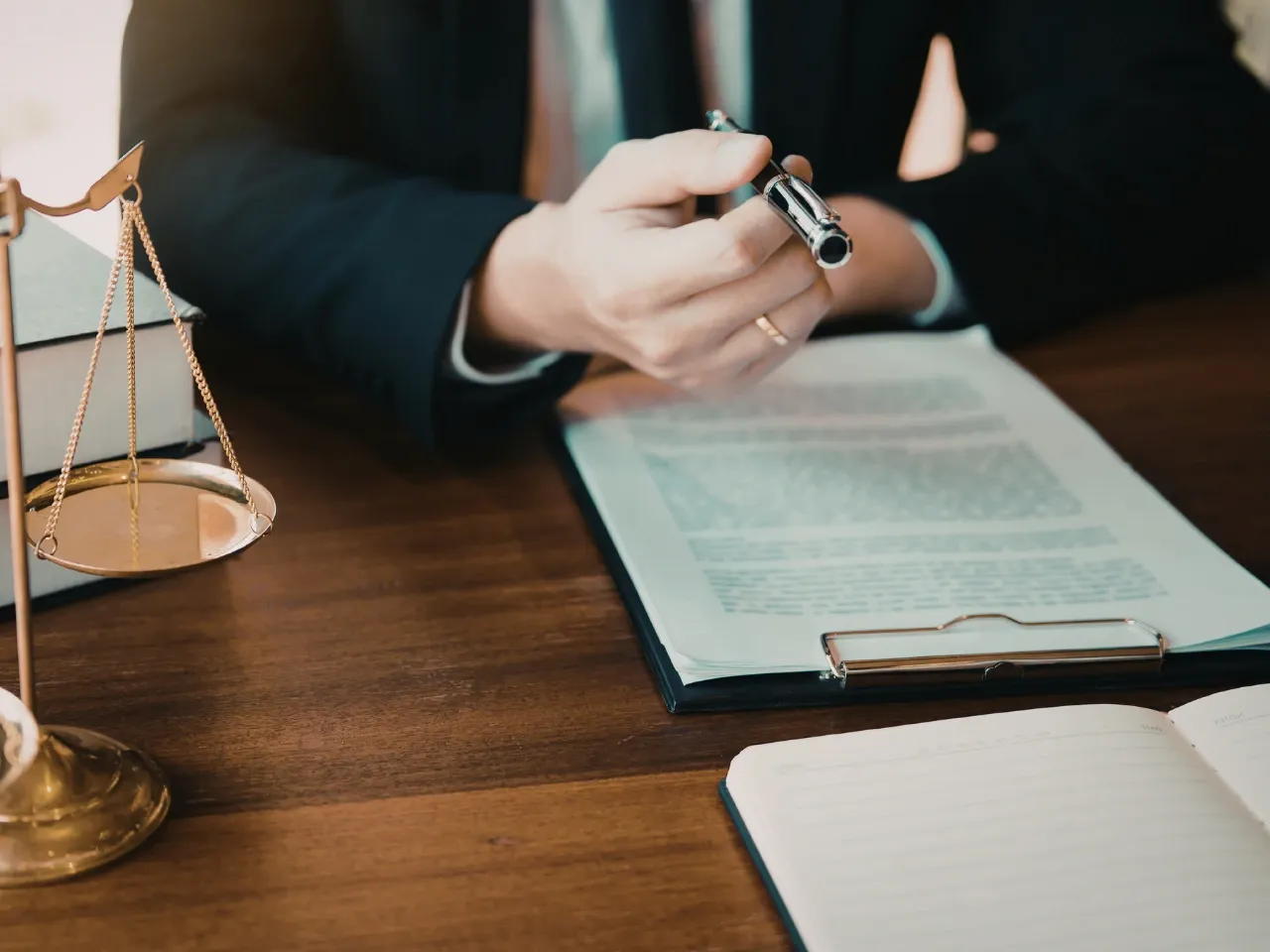 A person in a suit holding a pen sits at a wooden desk with open documents, a clipboard, and a set of golden balance scales nearby, suggesting a legal or business setting.