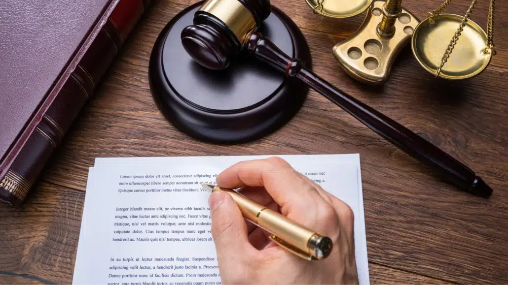 A person writes on a legal document at a desk with a gavel, a pair of scales, and a large book, suggesting a legal or judicial setting.