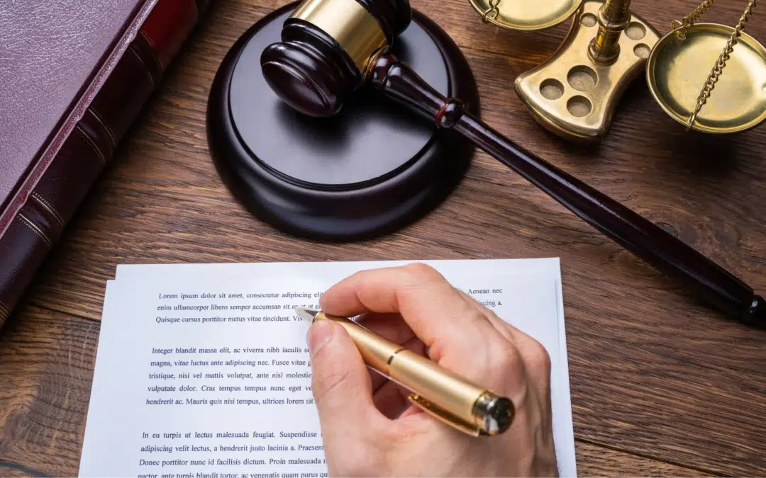 A person writes on a legal document at a desk with a gavel, a pair of scales, and a large book, suggesting a legal or judicial setting.