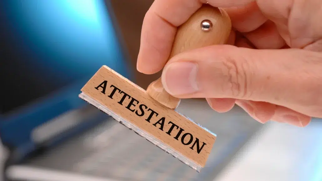 A hand holding a wooden stamp labeled ATTESTATION above a blurred laptop keyboard in the background.