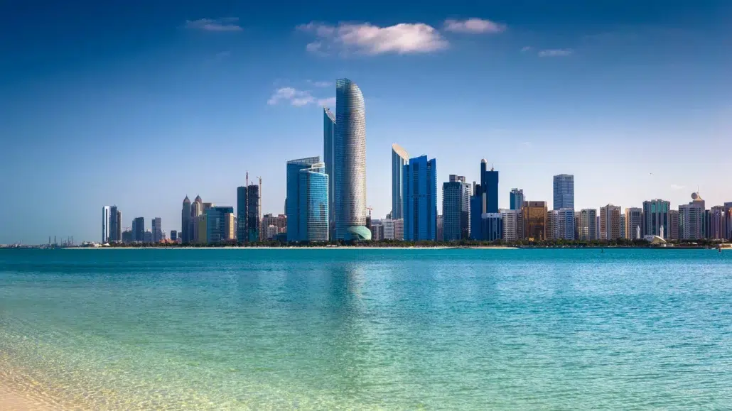 Abu Dhabi skyline with modern skyscrapers and high-rise buildings reflected in calm turquoise waters, set against a clear blue sky with a few clouds. Sandy shoreline is visible in the foreground.