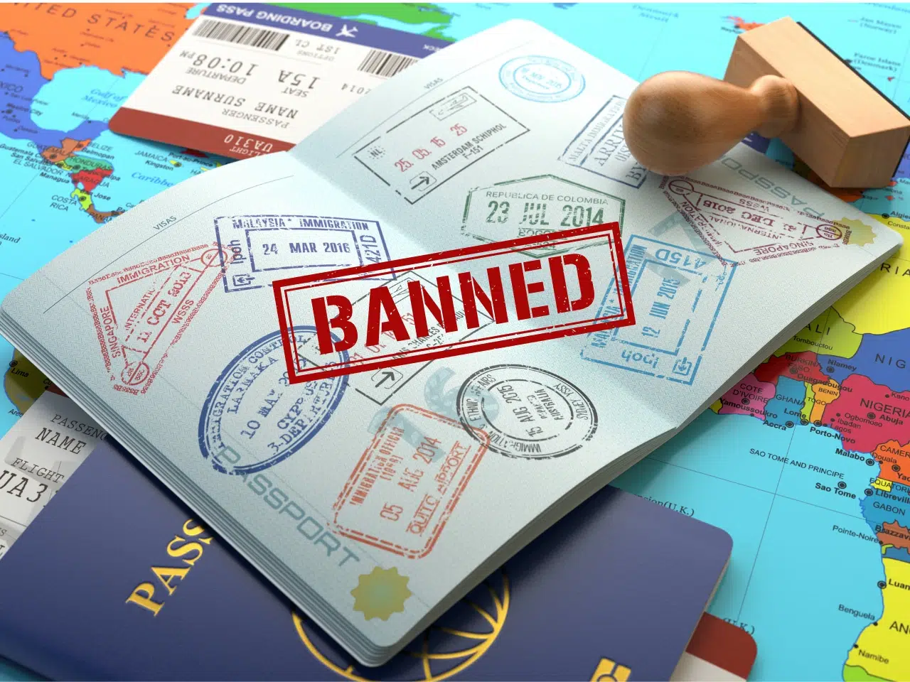 A passport with various entry stamps lies open on a world map, marked with a large red BANNED stamp. Nearby are another passport, airline tickets, and a wooden stamp.