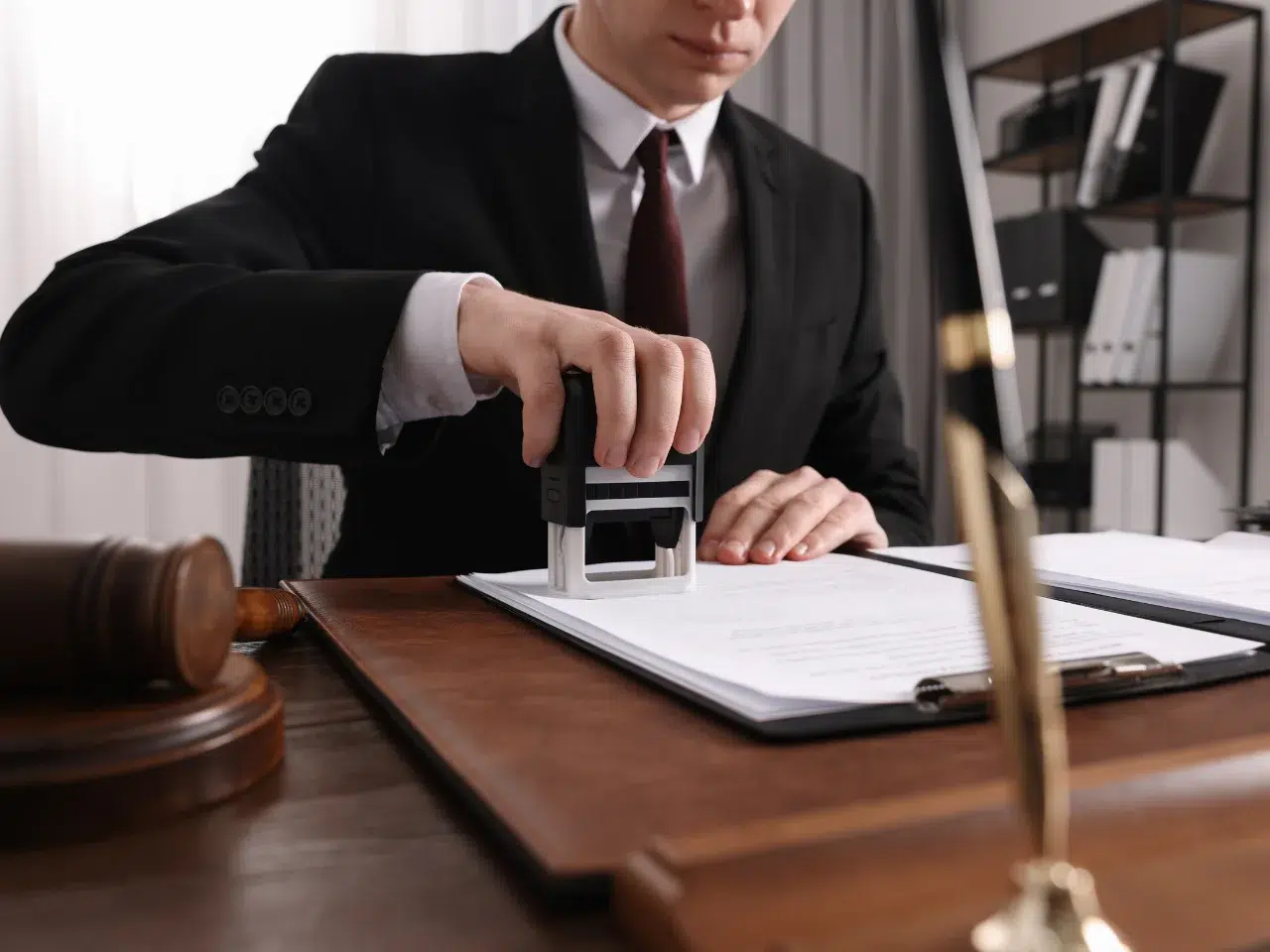 A person in a suit stamps a document at a desk with legal items, including a gavel and scales of justice, suggesting an official or legal setting.
