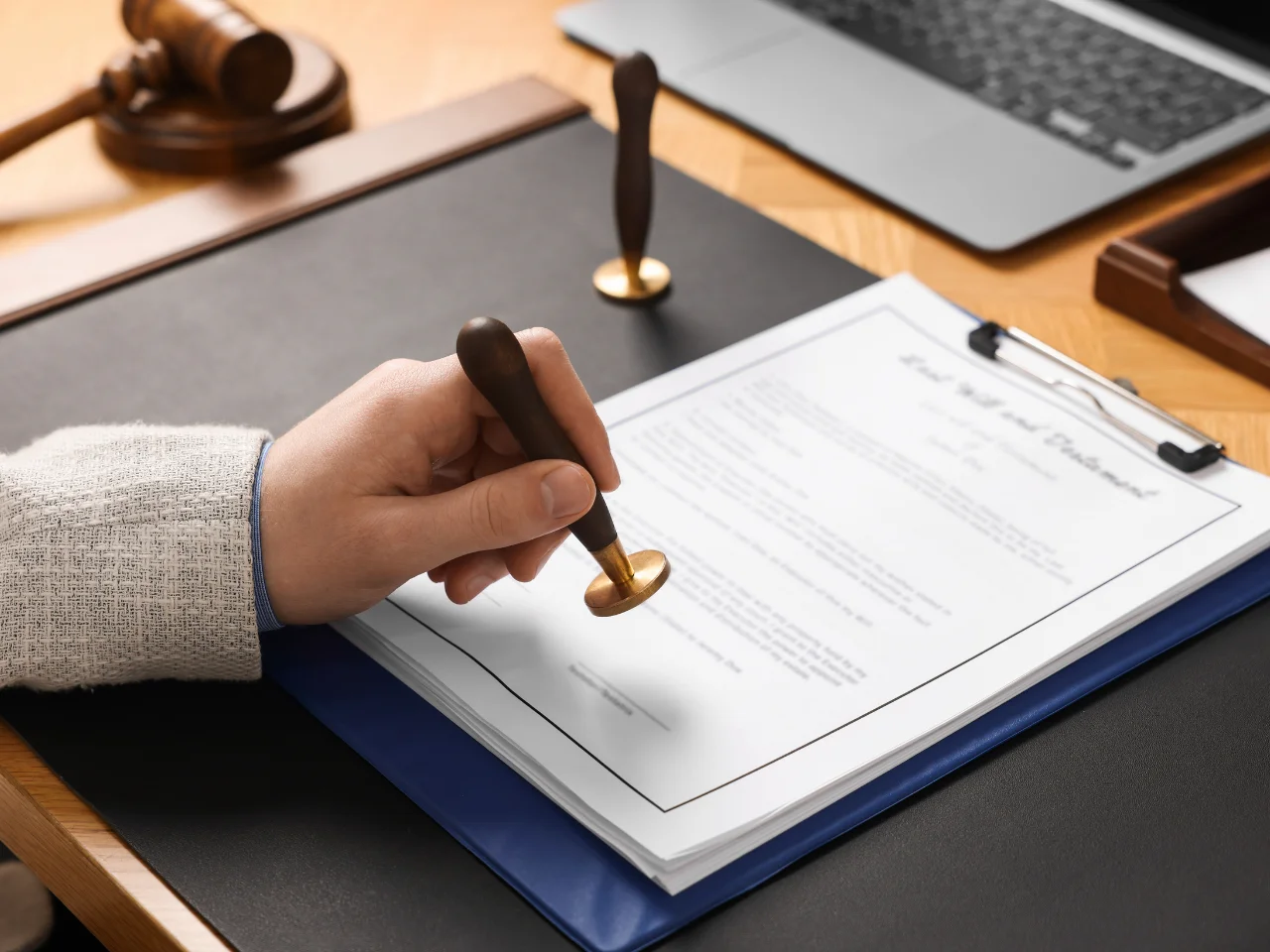A person stamps a document labeled Last Will and Testament on a desk with a gavel, a laptop, and another stamp visible nearby.