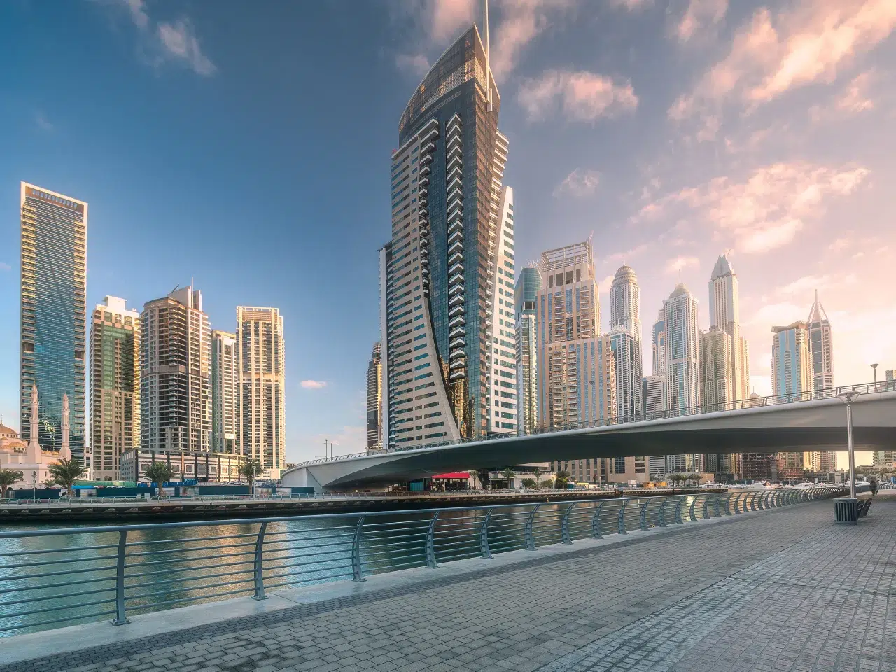 Modern skyscrapers line a waterfront in a cityscape at sunset, with a bridge spanning over the water and a wide pedestrian walkway with railings in the foreground under a partly cloudy sky.