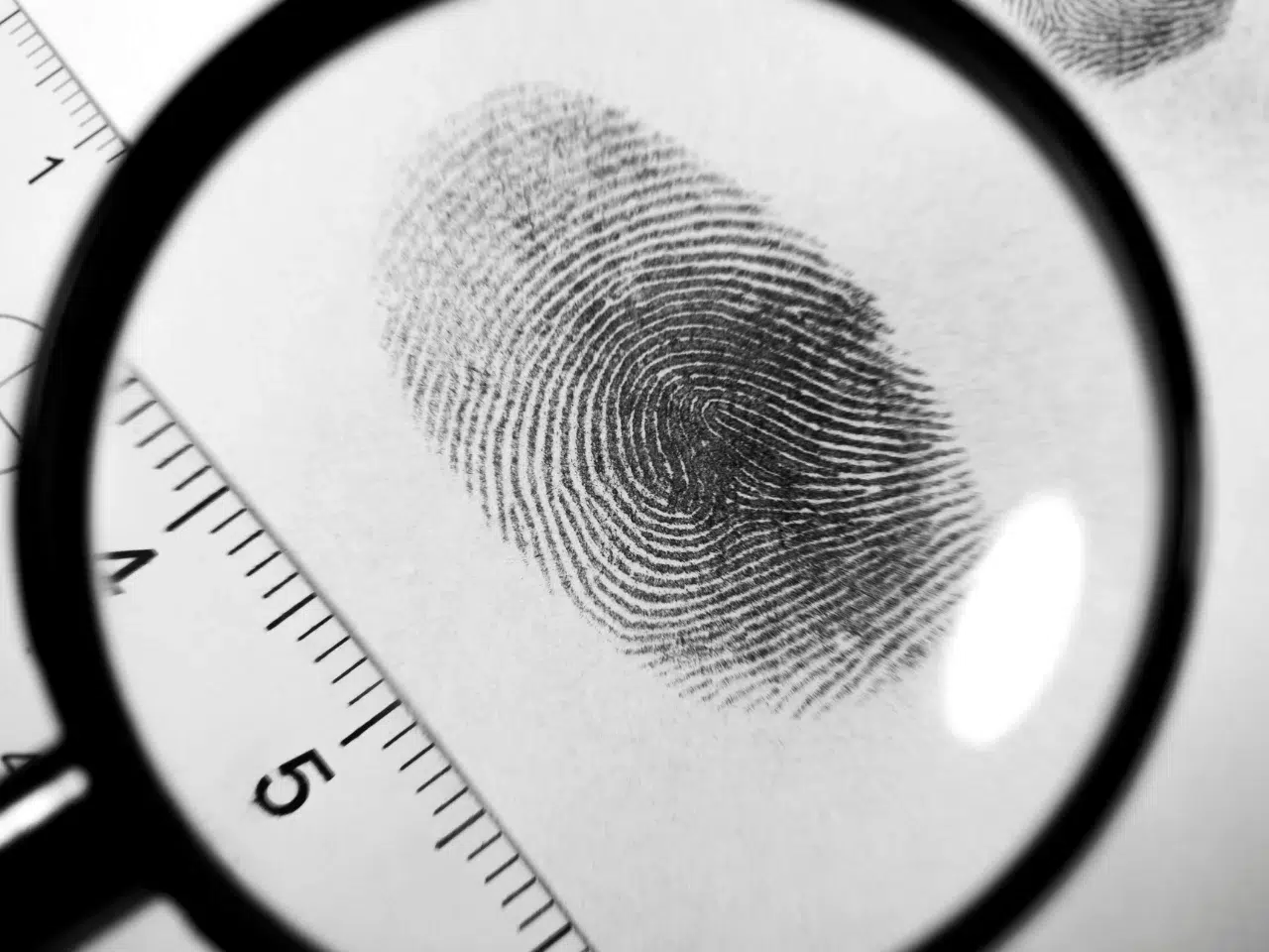 A black ink fingerprint is viewed through a magnifying glass, with a ruler beside it showing measurement marks. The image focuses on the fingerprint ridges and details.