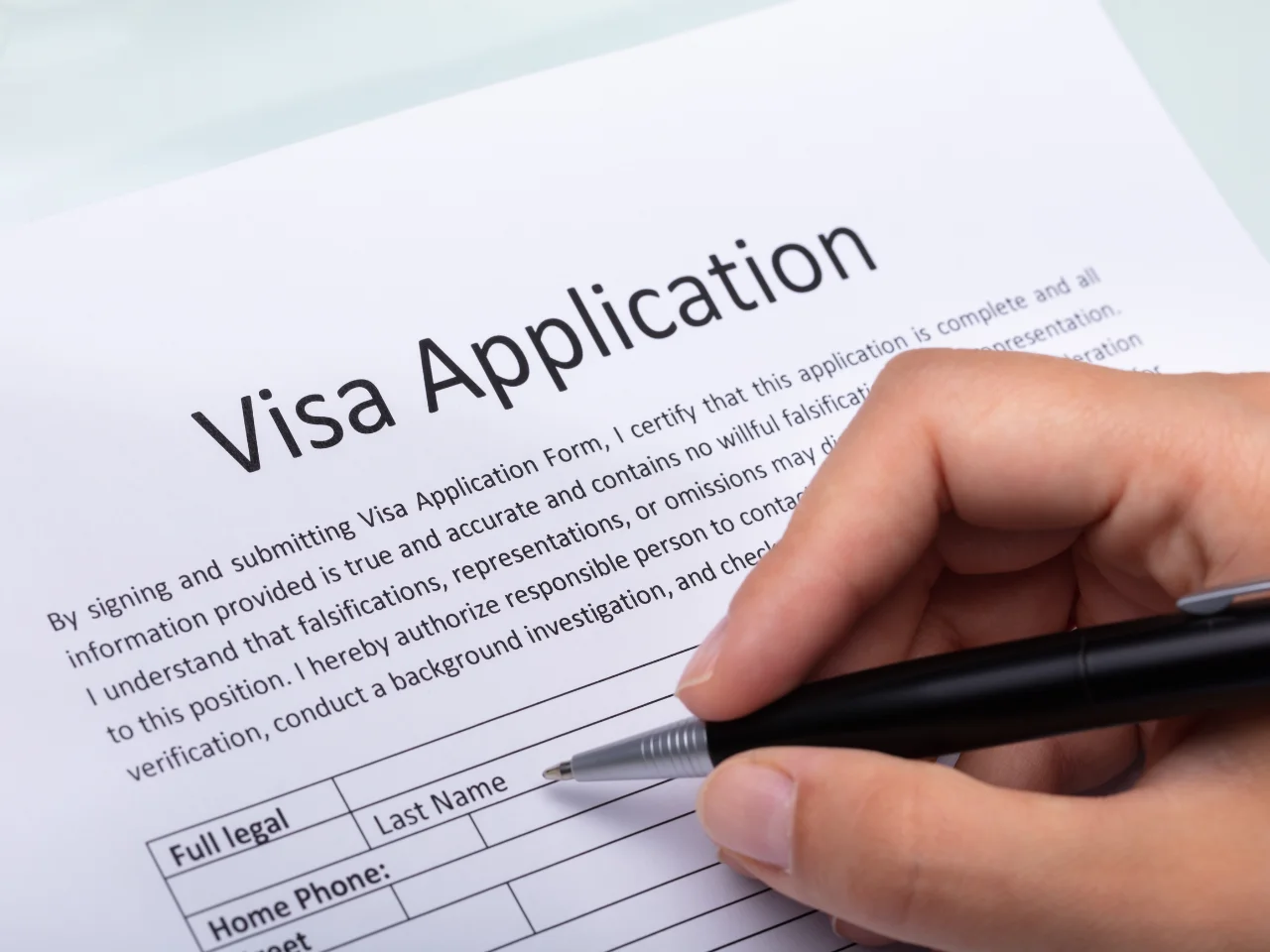 A hand holding a pen is filling out a visa application form. The section shown includes fields for full name and phone number. The top of the form displays the title Visa Application in bold letters.