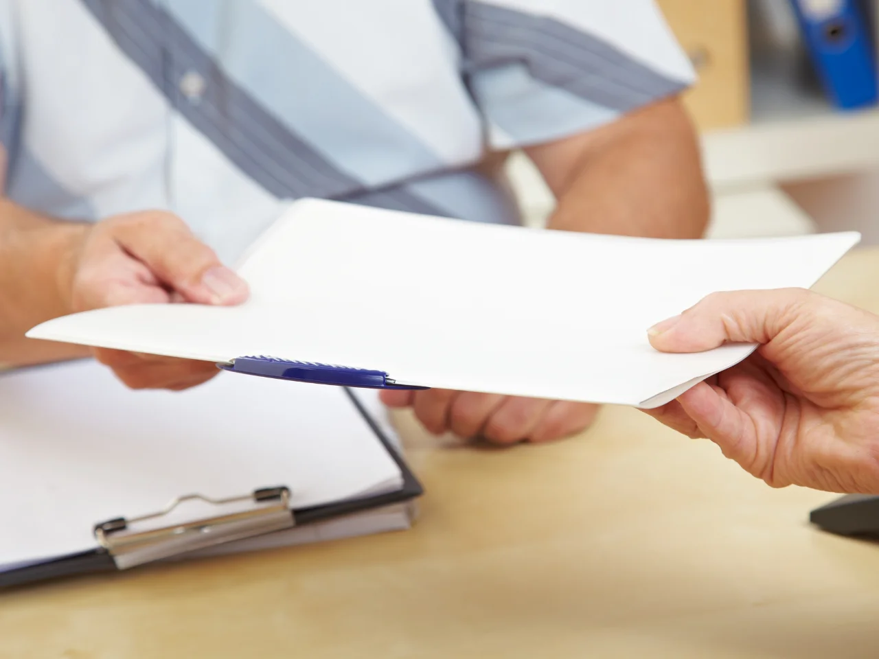 Two people exchanging a white folder over a desk; one person is handing it over while the other is receiving it. A clipboard with papers is on the desk in the background.