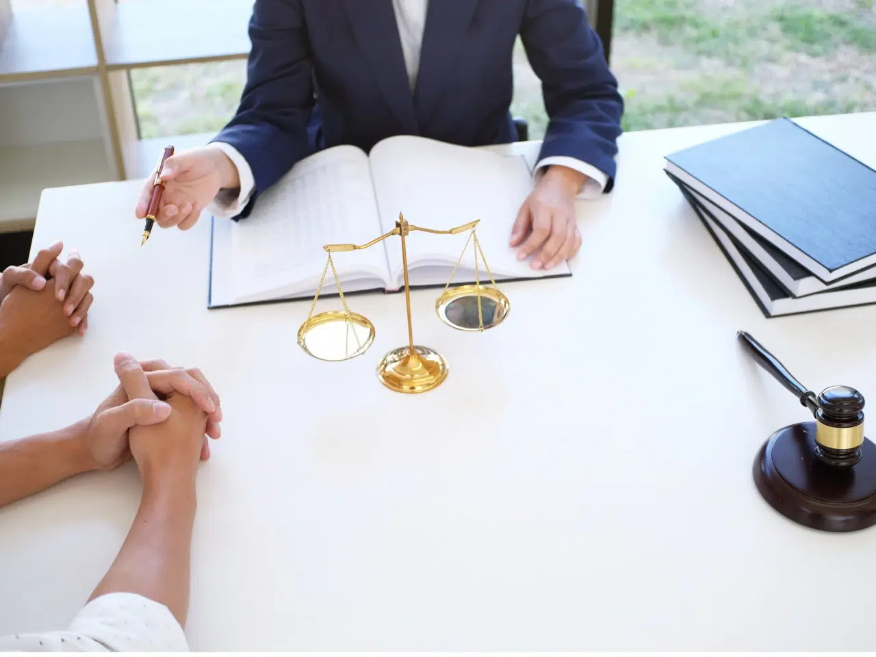 Three people sit at a white table with a scale of justice, gavel, laptop, and open book. One person in a suit gestures with a pen, suggesting a legal consultation or meeting.
