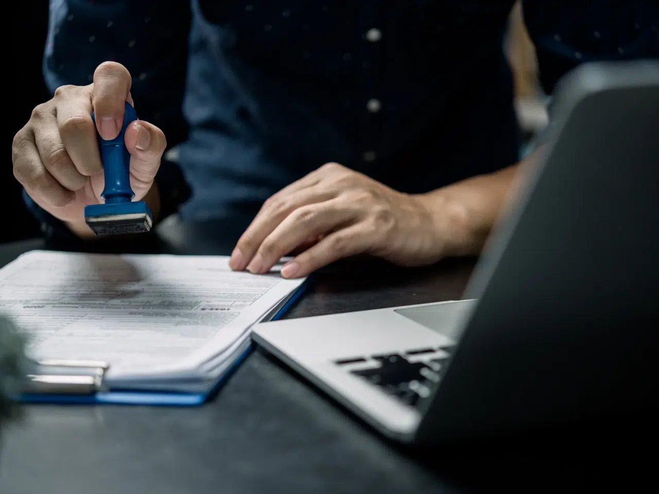 A person presses a blue stamp onto a document on a clipboard beside an open laptop, indicating approval or verification of paperwork.