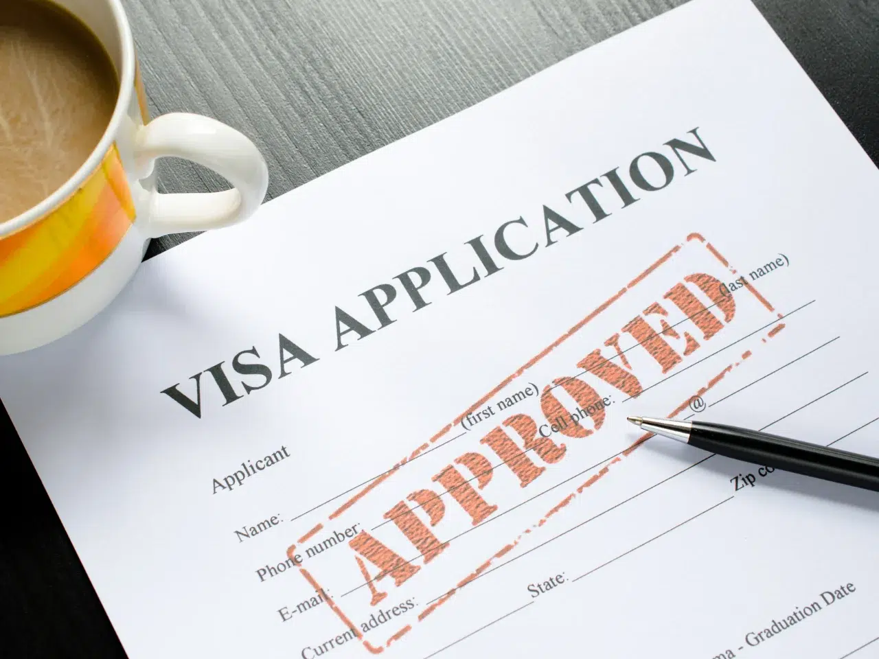 A visa application form with a large, red APPROVED stamp across it, a pen resting on the paper, and a cup of coffee beside it on a table.