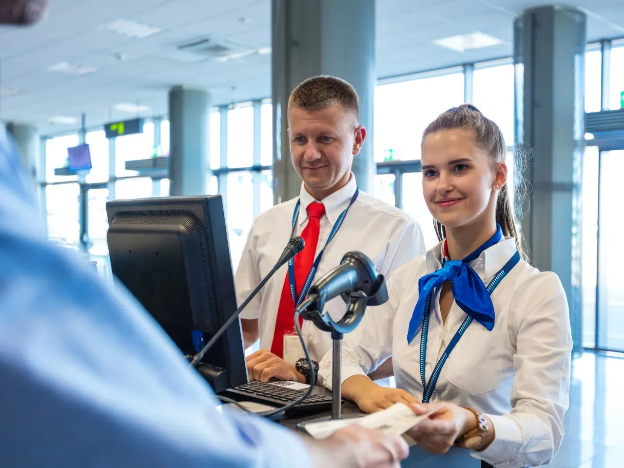 Two airport staff members in uniform assist a passenger at a check-in counter. One staff member scans a document while the other looks on. They are standing behind a computer and ticket scanner.