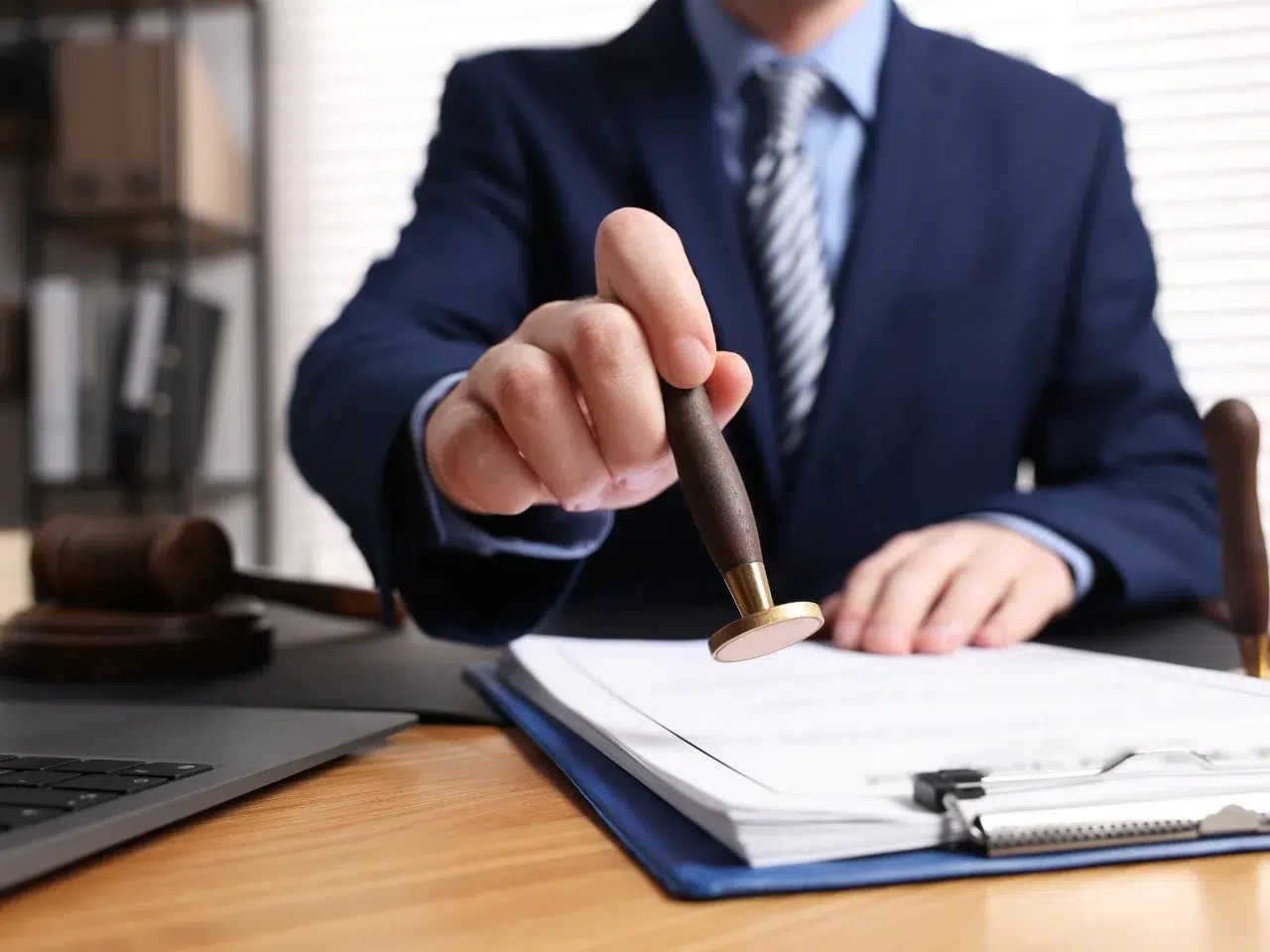 A person in a suit stamps a document on a desk, with a gavel, pen, and laptop nearby, suggesting a legal or official setting.