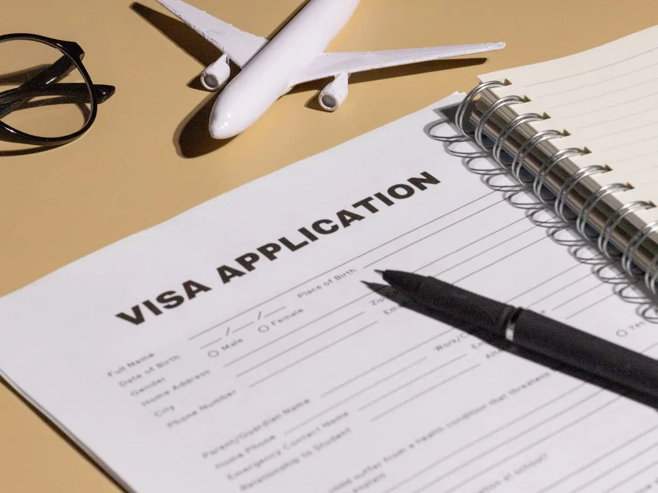 A visa application form on a desk with a black pen, a spiral notebook, eyeglasses, and a small model airplane, suggesting travel planning or preparation.