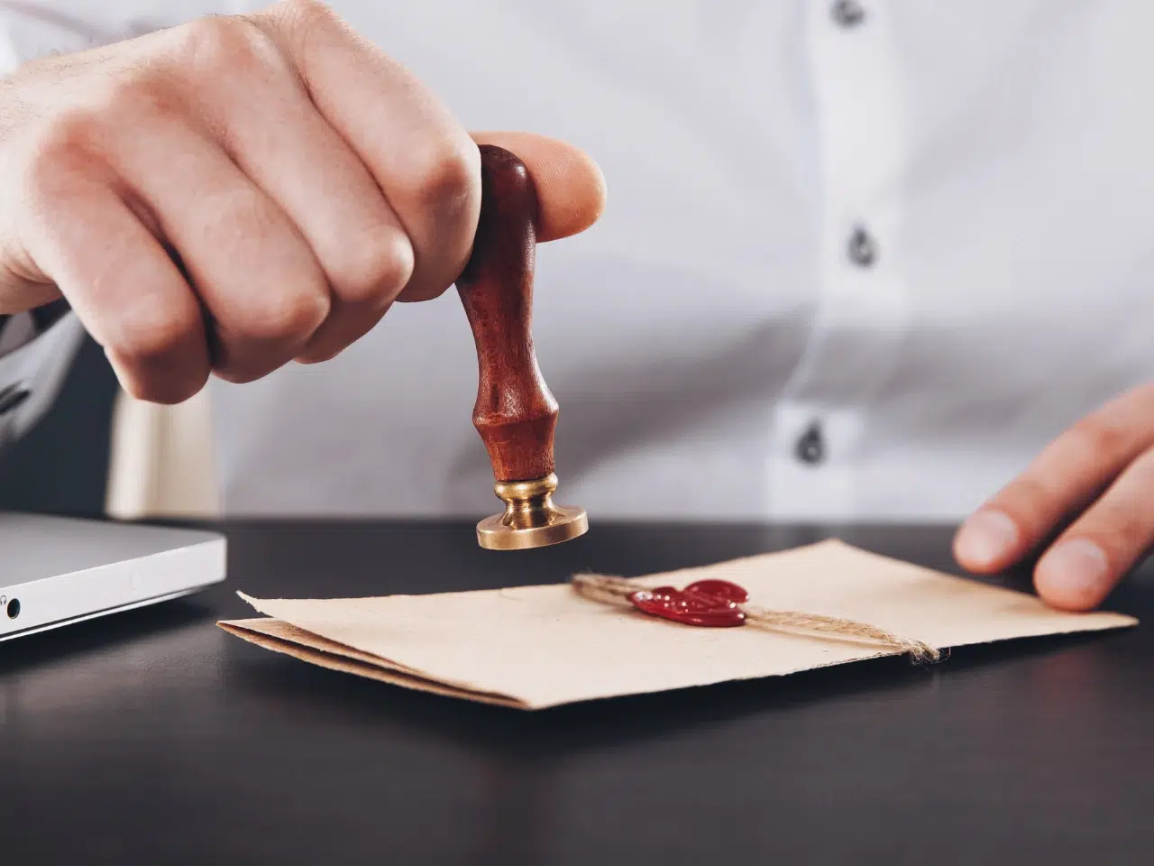 A person presses a wax seal stamp onto a tied envelope, sealing it with red wax. The person wears a white shirt, and the scene is set on a dark table near a laptop.