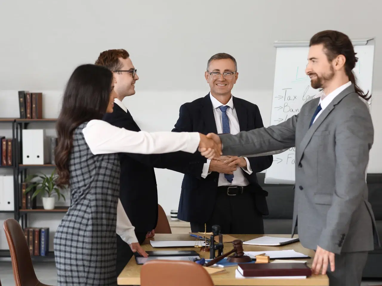 Four business professionals in formal attire are in an office. Two are shaking hands across a table with documents, while another man stands clapping and one woman looks on, all smiling. A whiteboard is visible in the background.