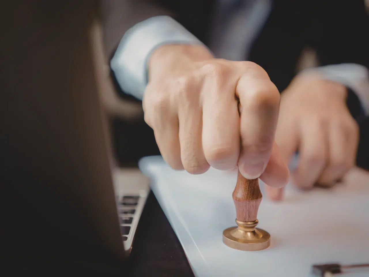 A close-up of a hand pressing a wooden stamp onto a document, with part of a laptop visible in the foreground, suggesting an official approval or authentication process.
