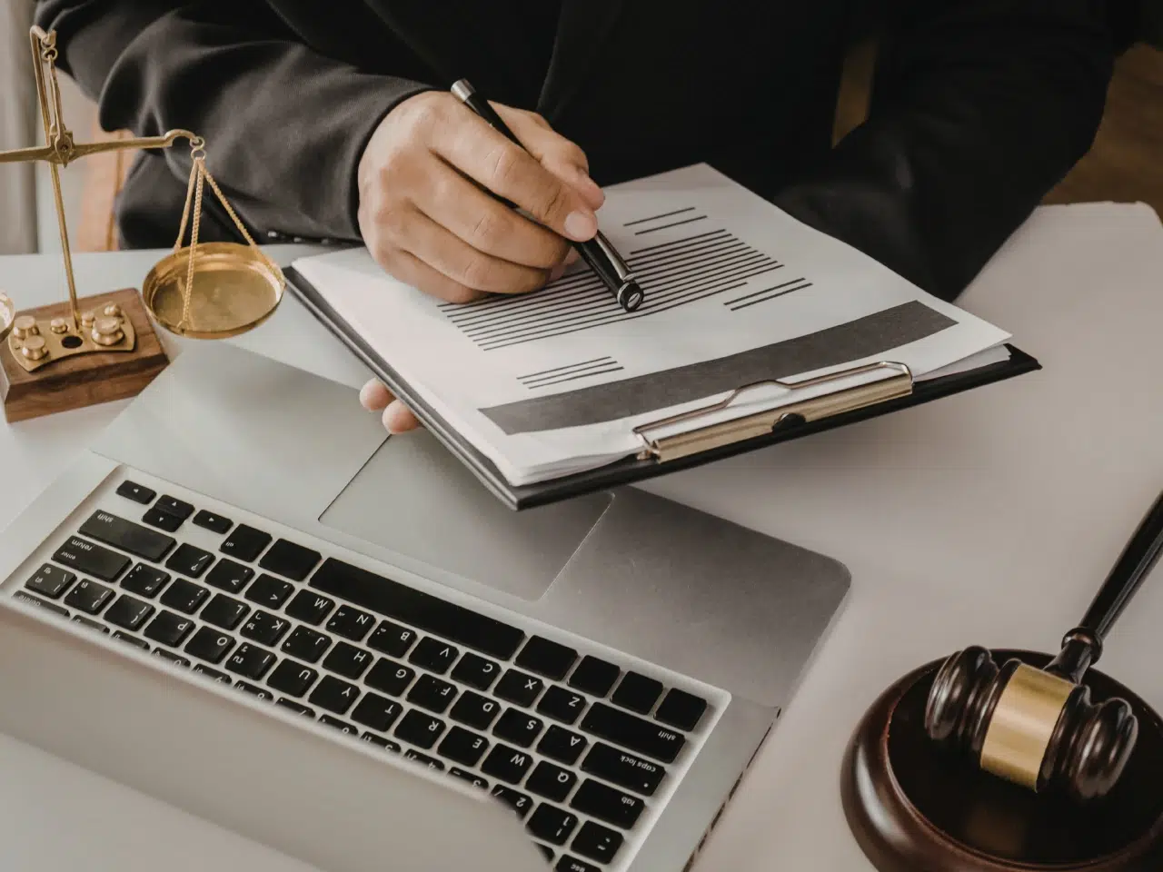 A person in a suit reviews a document on a clipboard beside a laptop, a judge’s gavel, and scales of justice on a desk, symbolizing legal or judicial work.