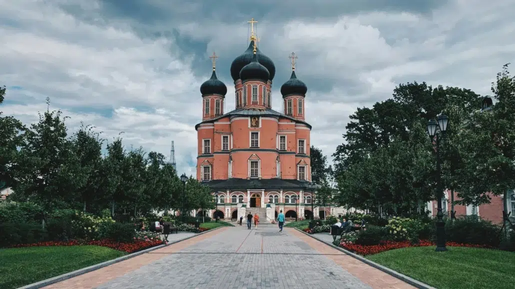 A view of a large red-brick Orthodox church with black onion domes and golden crosses, surrounded by trees and gardens, with people walking along a paved path under a cloudy sky.